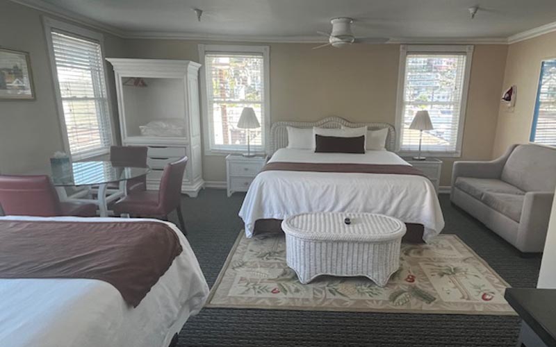 Spacious hotel room with a centered white bed, a second bed to the left, wicker coffee table, and a beige sofa by large windows.