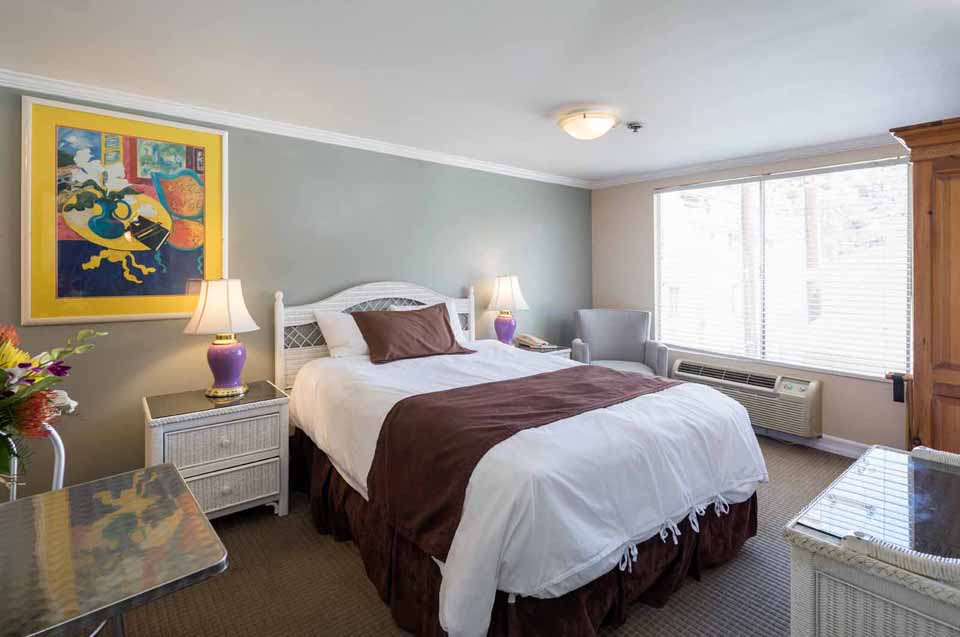 Hotel room with a white bed, brown runner, two nightstands, purple lamps, and a framed colorful painting on a gray wall side.